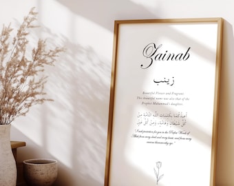 May include: A framed print with the name "Zainab" in elegant script. The print includes Arabic text and English translations, with a floral illustration at the bottom. The frame is a warm, light brown color.