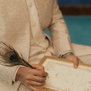 Puede incluir: Una chaqueta sherwani beige texturizada con cuello alto y botones, combinada con pantalones a juego. Una pluma de pavo real se sostiene sobre un documento rectangular en un marco dorado, con bordes florales y texto impreso.