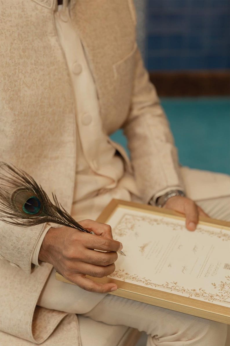 Puede incluir: Una chaqueta sherwani beige texturizada con cuello alto y botones, combinada con pantalones a juego. Una pluma de pavo real se sostiene sobre un documento rectangular en un marco dorado, con bordes florales y texto impreso.