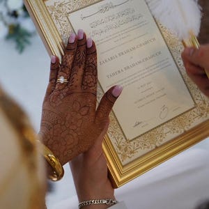 Może przedstawiać: Złota rama z aktem małżeństwa ozdobiona misternymi dekoracyjnymi obramowaniami i arabską kaligrafią u g&oacute;ry. Dokument zawiera nazwiska Zakaria Braham-Chaouch i Tahera Braham-Chaouch, datowany na 30 maja 2017 roku, wraz z białym pi&oacute;rem do pisania.