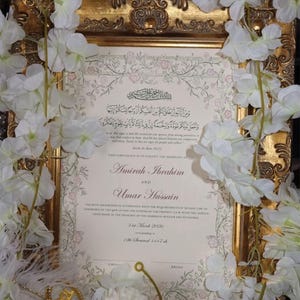 May include: A marriage certificate for Amirah Ibrahim and Umar Hussain, dated 31st March 2026, displayed in an ornate gold frame. The document features Arabic calligraphy, English text, and a floral border, surrounded by white artificial flowers.