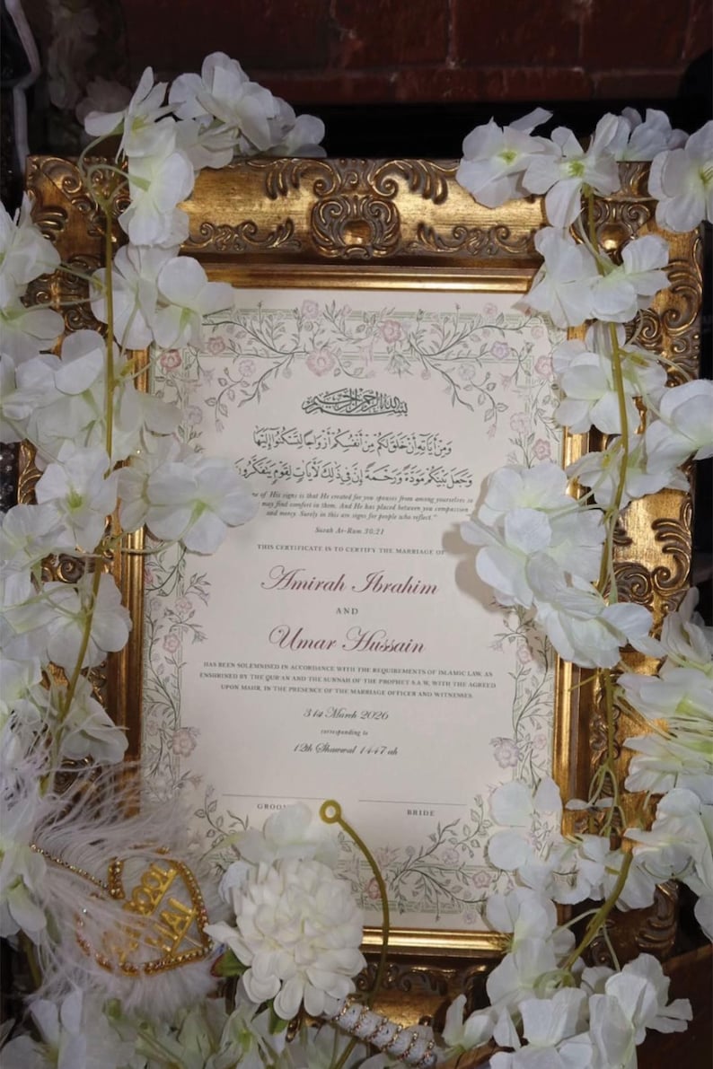 May include: A marriage certificate for Amirah Ibrahim and Umar Hussain, dated 31st March 2026, displayed in an ornate gold frame. The document features Arabic calligraphy, English text, and a floral border, surrounded by white artificial flowers.