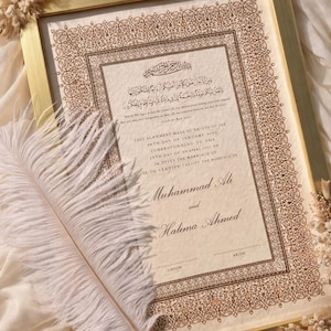 K&ouml;nnte beinhalten: Eine gerahmte Heiratsurkunde mit goldfarbenem Rahmen, verziert mit komplexen braunen Blumenmustern, arabischer Kalligrafie und englischem Text mit den Namen Muhammad Ali und Halima Ahmed, erg&auml;nzt durch eine wei&szlig;e Strau&szlig;enfeder als Schreibfeder.