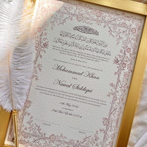 Könnte beinhalten: Eine gerahmte islamische Heiratsurkunde mit goldenem Rand und floralen Mustern in gedämpfter roter Tinte. Das Dokument enthält arabische Kalligrafie, englischen Text, die Namen Mohammad Khan und Nawal Siddiqui sowie einen weißen Federkiel.
