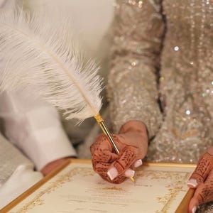Puede incluir: Una pluma blanca con punta de metal dorado utilizada para firmar un documento enmarcado en oro. El documento presenta bordes decorativos dorados intrincados y texto impreso, sostenido por manos adornadas con detallados patrones de henna marr&oacute;n.
