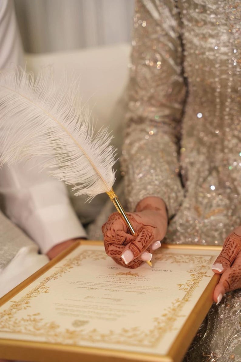 Puede incluir: Una pluma blanca con punta de metal dorado utilizada para firmar un documento enmarcado en oro. El documento presenta bordes decorativos dorados intrincados y texto impreso, sostenido por manos adornadas con detallados patrones de henna marr&oacute;n.