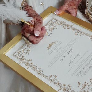 Puede incluir: Un certificado de matrimonio enmarcado con bordes decorativos dorados y el texto Naeem Syed y Mahha Malik, fechado el 22 de mayo de 2025. El documento es sostenido por manos con patrones de henna y una pluma blanca descansa sobre el papel.