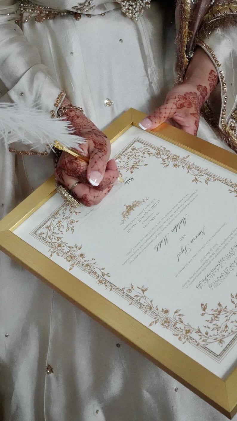Puede incluir: Un certificado de matrimonio enmarcado con bordes decorativos dorados y el texto Naeem Syed y Mahha Malik, fechado el 22 de mayo de 2025. El documento es sostenido por manos con patrones de henna y una pluma blanca descansa sobre el papel.