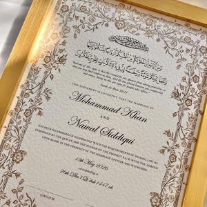 Könnte beinhalten: Eine gerahmte islamische Heiratsurkunde mit goldenen Metallrändern. Das Dokument zeigt komplizierte florale Illustrationen in brauner Tinte, arabische Kalligrafie und englischen Text mit den Namen Mohammad Khan und Nawal Siddiqui, datiert auf den 13. Mai 2026.