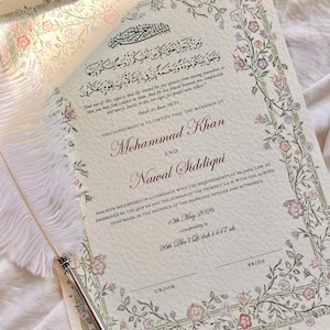 May include: A marriage certificate document featuring Arabic calligraphy, English text, and a floral border in shades of pink, purple, and green. A white feather quill pen with a silver metal tip rests diagonally across the paper surface.
