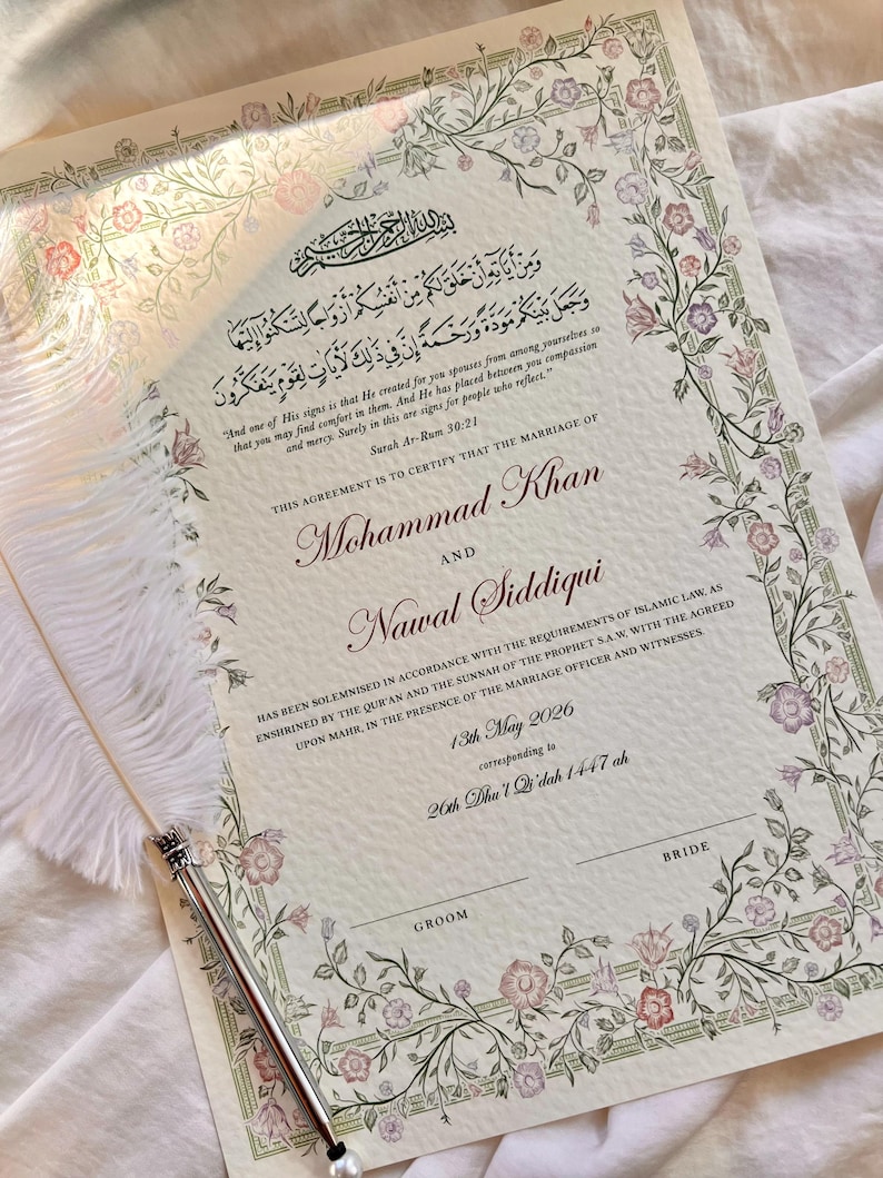 May include: A marriage certificate document featuring Arabic calligraphy, English text, and a floral border in shades of pink, purple, and green. A white feather quill pen with a silver metal tip rests diagonally across the paper surface.