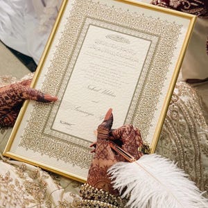 K&ouml;nnte beinhalten: Eine rechteckige Heiratsurkunde in einem goldenen Rahmen mit komplizierten goldenen Randmustern. Das Dokument enth&auml;lt gedruckten Text in Englisch und arabischer Schrift, einschlie&szlig;lich der Namen Nahid Uddin und Sumaiya A. Eine wei&szlig;e Federkiel-Stift ist vorhanden.