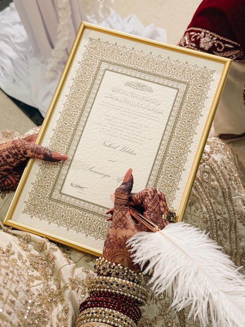 K&ouml;nnte beinhalten: Eine rechteckige Heiratsurkunde in einem goldenen Rahmen mit komplizierten goldenen Randmustern. Das Dokument enth&auml;lt gedruckten Text in Englisch und arabischer Schrift, einschlie&szlig;lich der Namen Nahid Uddin und Sumaiya A. Eine wei&szlig;e Federkiel-Stift ist vorhanden.