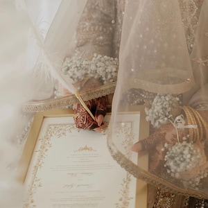 Puede incluir: Un documento con marco dorado y bordes florales intrincados que se firma con una pluma blanca. El documento contiene texto impreso y es sostenido por manos adornadas con patrones de henna, brazaletes dorados y pulseras de flores blancas.