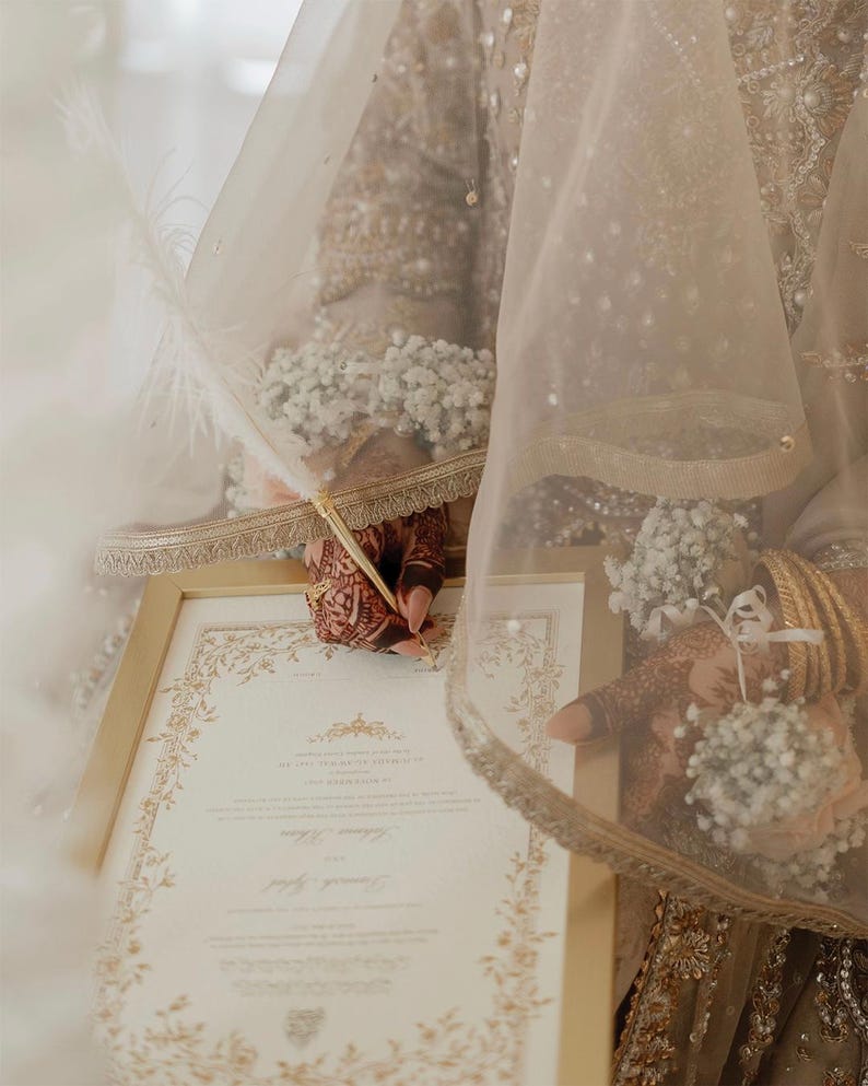 Puede incluir: Un documento con marco dorado y bordes florales intrincados que se firma con una pluma blanca. El documento contiene texto impreso y es sostenido por manos adornadas con patrones de henna, brazaletes dorados y pulseras de flores blancas.