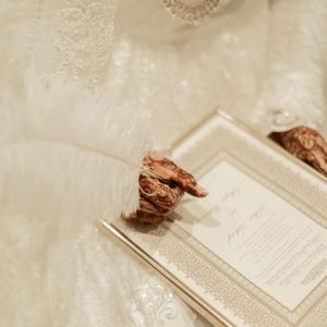 K&ouml;nnte beinhalten: Ein wei&szlig;es Spitzenkleidungsst&uuml;ck mit aufwendigen Stickereien und einer langen silbernen Halskette. Eine wei&szlig;e Feder liegt auf einem rechteckigen Dokument mit goldfarbenen R&auml;ndern und gedrucktem Text, gehalten von H&auml;nden mit braunen Hennamustern.