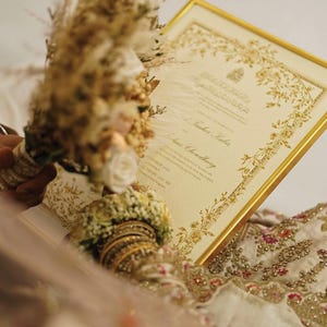Puede incluir: Un certificado rectangular en un marco dorado con bordes florales dorados intrincados y texto impreso. El certificado se sostiene junto a un ramo de flores beige secas y rosas blancas, con un vistazo a una prenda de tela color crema bordada.