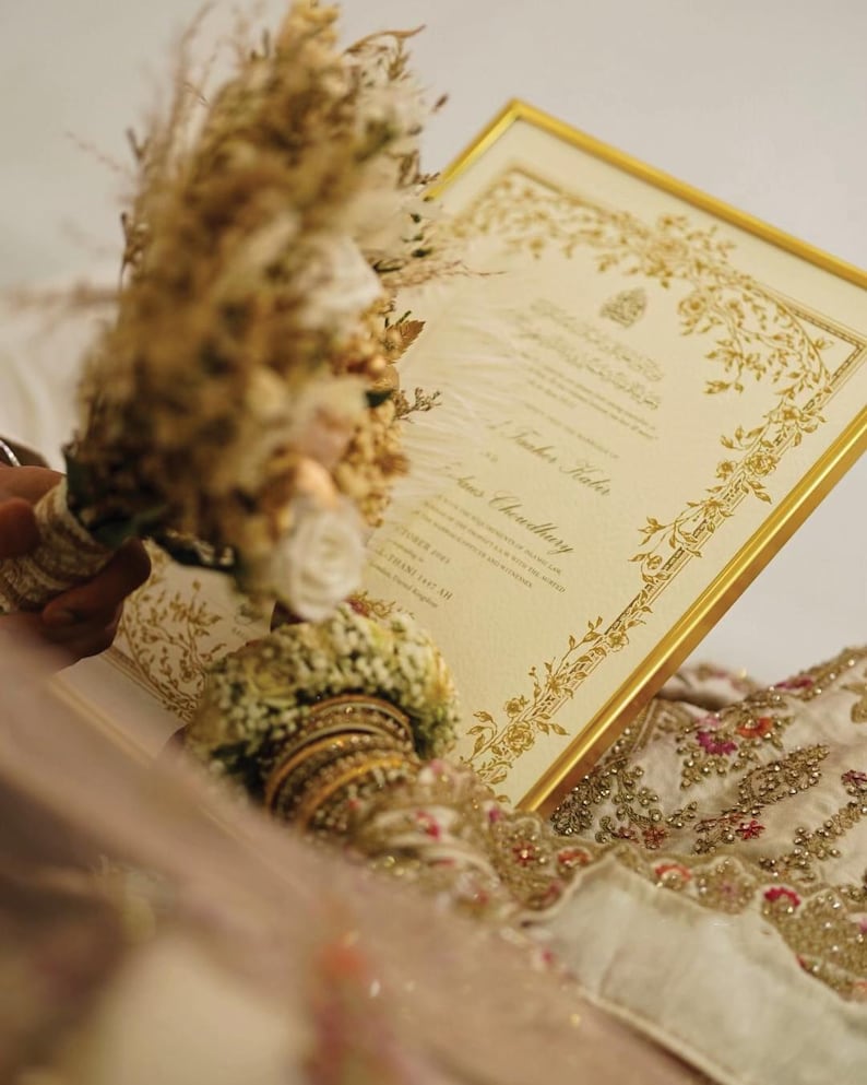Puede incluir: Un certificado rectangular en un marco dorado con bordes florales dorados intrincados y texto impreso. El certificado se sostiene junto a un ramo de flores beige secas y rosas blancas, con un vistazo a una prenda de tela color crema bordada.