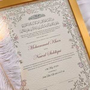 May include: A framed Islamic marriage certificate with a gold border, featuring floral illustrations in pink and green. The document includes Arabic calligraphy, English text, the names Mohammad Khan and Nawal Siddiqui, and a white feather quill pen.