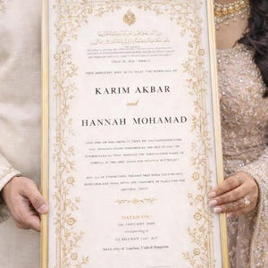 Puede incluir: Un certificado de matrimonio enmarcado con detalles florales dorados, sostenido por una pareja. El documento presenta los nombres de Karim Akbar y Hannah Mohamad, con texto en inglés y árabe. Con fecha de enero de 2006.