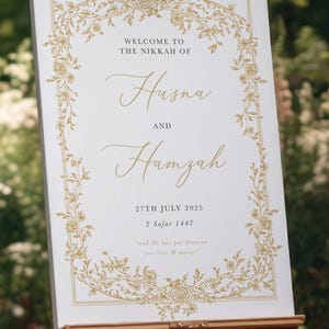 May include: A white wedding sign on an easel with gold floral detailing. The sign reads "WELCOME TO THE NIKKAH OF Husna AND Hamzah" with the date "27TH JULY 2025" and a quote. The sign is displayed outdoors.