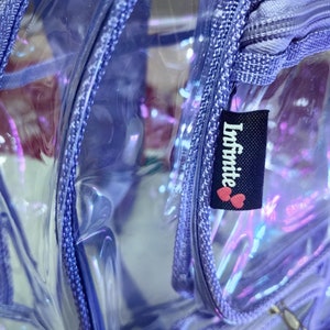 Op de afbeelding: Een transparante, paarse regenboogkleurige rugzak met een ritssluiting en een zwart label met de tekst "Infinite" met twee roze harten.