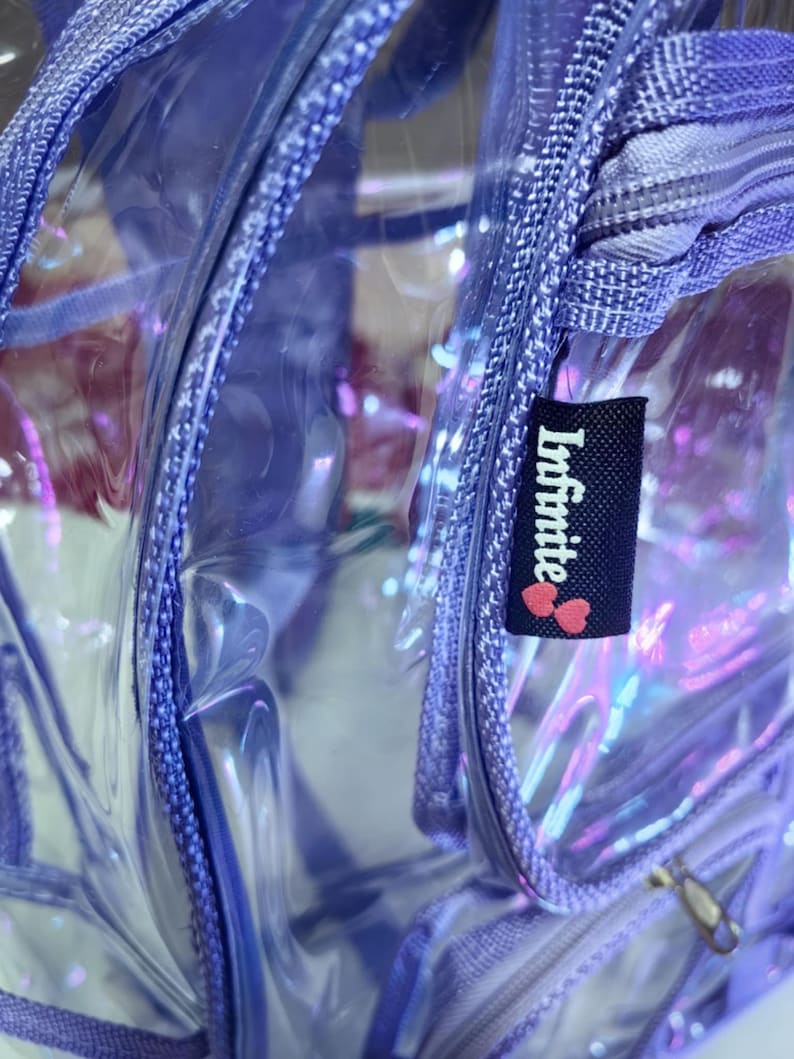 Op de afbeelding: Een transparante, paarse regenboogkleurige rugzak met een ritssluiting en een zwart label met de tekst "Infinite" met twee roze harten.
