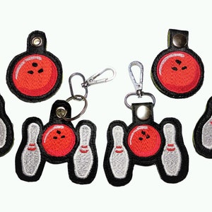 May include: Set of six embroidered keychains featuring bowling pins and balls. The keychains are black with red and white stitching. The bowling pins are white with red stripes. The bowling balls are red with black stitching.