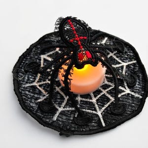 May include: A circular black lace doily featuring a white embroidered spiderweb pattern and a three-dimensional black lace spider with red stitching accents. A small, round, battery-operated LED candle with a warm orange glow sits in the center.
