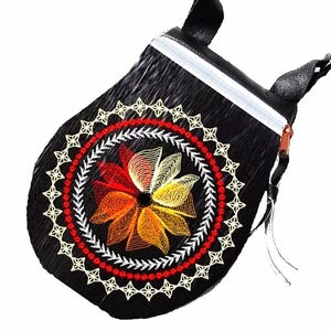 May include: Black crossbody bag with a floral embroidery design. The bag features a colorful flower in the center, surrounded by a red and white border. It has a white zipper and a brown strap.