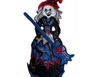 May include: An embroidered patch featuring a witch with gray hair, a red hat, and a blue dress. The witch holds a blue broomstick with red accents. The design includes black, white, blue, red, and green thread colors on a fabric base.