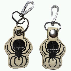 May include: Two beige leather keyrings with a black embroidered spider design. One keyring has a silver split ring and a silver lobster clasp. The other keyring has a silver lobster clasp and a silver button.