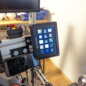 Könnte beinhalten: Ein schwarzes Stream Deck mit einem Raster aus 12 Tasten, jede mit einem anderen Symbol. Die Tasten sind mit blauem Licht beleuchtet. Das Stream Deck ist an einer silbernen Metallstange montiert.