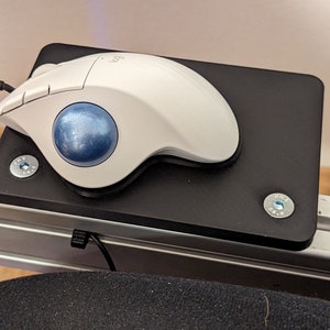 Könnte beinhalten: Eine weiße ergonomische Trackball-Maus mit blauem Trackball liegt auf einer schwarzen rechteckigen Plattform. Die Plattform ist mit silbernen Schrauben an einem Metallrahmen befestigt. Die Maus ist über ein schwarzes Kabel verbunden.