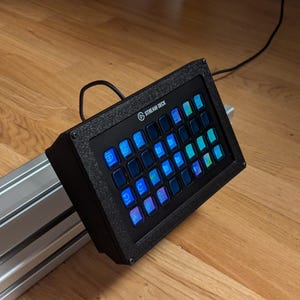 Könnte beinhalten: Ein schwarzes Stream Deck mit einem Raster aus 32 anpassbaren Tasten, jede mit einem anderen farbigen Licht. Das Stream Deck ist auf einer silbernen Metallschiene montiert.