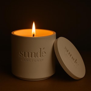 Pode incluir: Uma vela bege acesa num recipiente redondo com tampa. A chama brilha intensamente contra o fundo escuro. O recipiente tem as palavras "sundé" e "SANCTUARY" em relevo. A tampa também tem a palavra "sundé" em relevo.