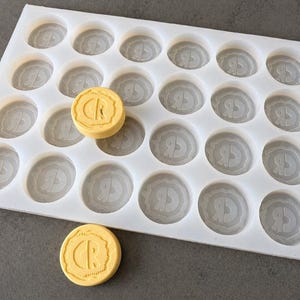May include: A white silicone mold with 24 circular cavities, each with a detailed design. Two yellow, circular soaps with the letters "CR" are visible. The mold is designed for creating soaps or similar crafts.
