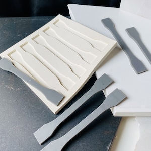 May include: A white rectangular mold with six cavities, each shaped like a dumbbell. Several gray dumbbell-shaped objects are scattered around the mold. The objects and mold are on a gray and white surface.