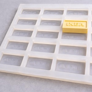 Puede incluir: Un molde de silicona blanco con compartimentos rectangulares, cada uno con el logotipo de IKEA. Un solo jabón rectangular amarillo con el texto IKEA se coloca en uno de los compartimentos. El molde está diseñado para hacer jabón u otros artículos pequeños.