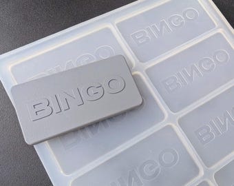 May include: A gray silicone mold with six rectangular cavities. Each cavity has the word "BINGO" embossed in it. A gray plastic card with the word "BINGO" embossed on it is sitting on top of the mold.