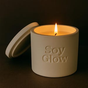Könnte beinhalten: Eine brennende Sojakerze in einem cremefarbenen Keramikbehälter mit dem geprägten Text "Soy Glow". Der Deckel liegt daneben. Die Kerzenflamme leuchtet vor dunklem Hintergrund.