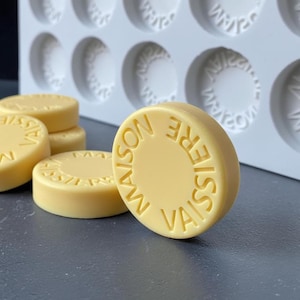 May include: Several pale yellow, circular wax seals or soap discs featuring the embossed text MAISON VAISSIERE arranged in a circle. A light gray silicone mold with multiple circular cavities is positioned in the background behind the discs.