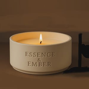 Pode incluir: Uma vela acesa, de cor creme, num recipiente redondo de cerâmica. A superfície da vela é lisa e a chama é brilhante. As palavras "ESSENCE & EMBER" estão gravadas na lateral do recipiente. Um artigo de decoração para casa.