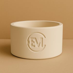 Könnte beinhalten: Ein cremefarbener, zylindrischer Pflanztopf mit einem geprägten "EM"-Logo. Der Pflanztopf hat eine breite Öffnung und eine glatte, matte Oberfläche. Das Design ist minimalistisch und modern, geeignet für den Innen- oder Außenbereich.
