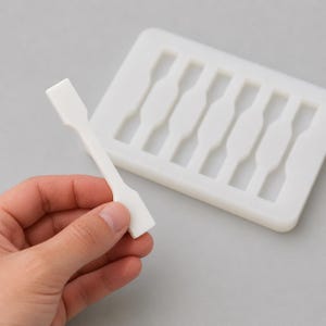 Könnte beinhalten: Eine weiße Kunststoffform mit sechs Hohlräumen, die jeweils wie eine kleine Hantel geformt sind. Ein passendes weißes, hantelförmiges Objekt wird in einer Hand gehalten, neben der Form. Die Objekte befinden sich auf einer hellgrauen Oberfläche.