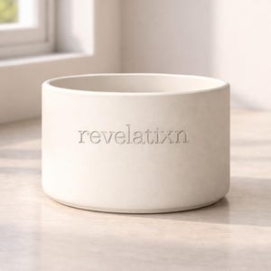 Puede incluir: Un cuenco cilíndrico de cerámica en color blanco roto con acabado mate, base plana y paredes rectas. La palabra "revelatixn" está grabada en relieve con una tipografía con serifa en el centro de la parte frontal. La superficie es uniforme.