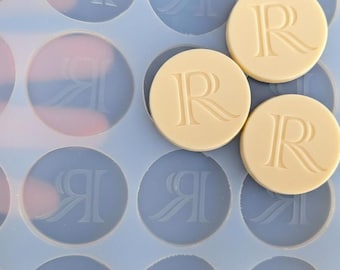 May include: A close-up shot of a light blue silicone mold with circular indentations. Three beige, circular objects with the letter "R" are visible. The mold has a repeating pattern of a stylized letter "R" in each indentation.