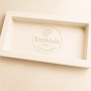 May include: An off-white rectangular tray with a raised border. The centre has a circular logo with the text "RotaMade HANDMADE" and a leaf design. The tray is likely for serving or display purposes.