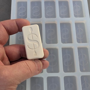 May include: A rectangular white wax or soap bar featuring an embossed dollar sign symbol with a vertical line through it. Behind the bar is a translucent silicone mold tray containing multiple identical rectangular cavities with the same symbol.