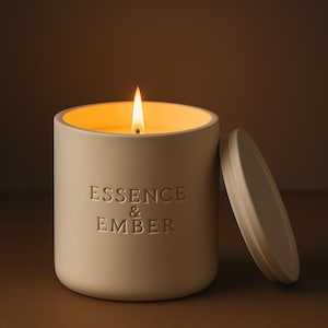May include: A lit, cream-colored candle in a ceramic container with the words "Essence & Ember" embossed on the side. The lid is off to the side. The candle's flame glows brightly against a warm background.